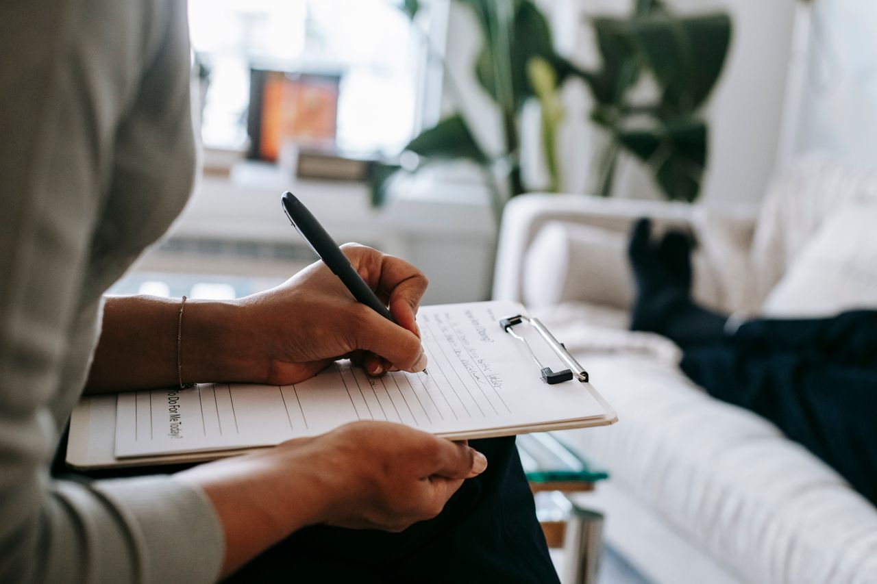 services-03 Unrecognizable ethnic female therapist taking notes on clipboard while filling out form during psychological appointment with anonymous client lying on blurred background