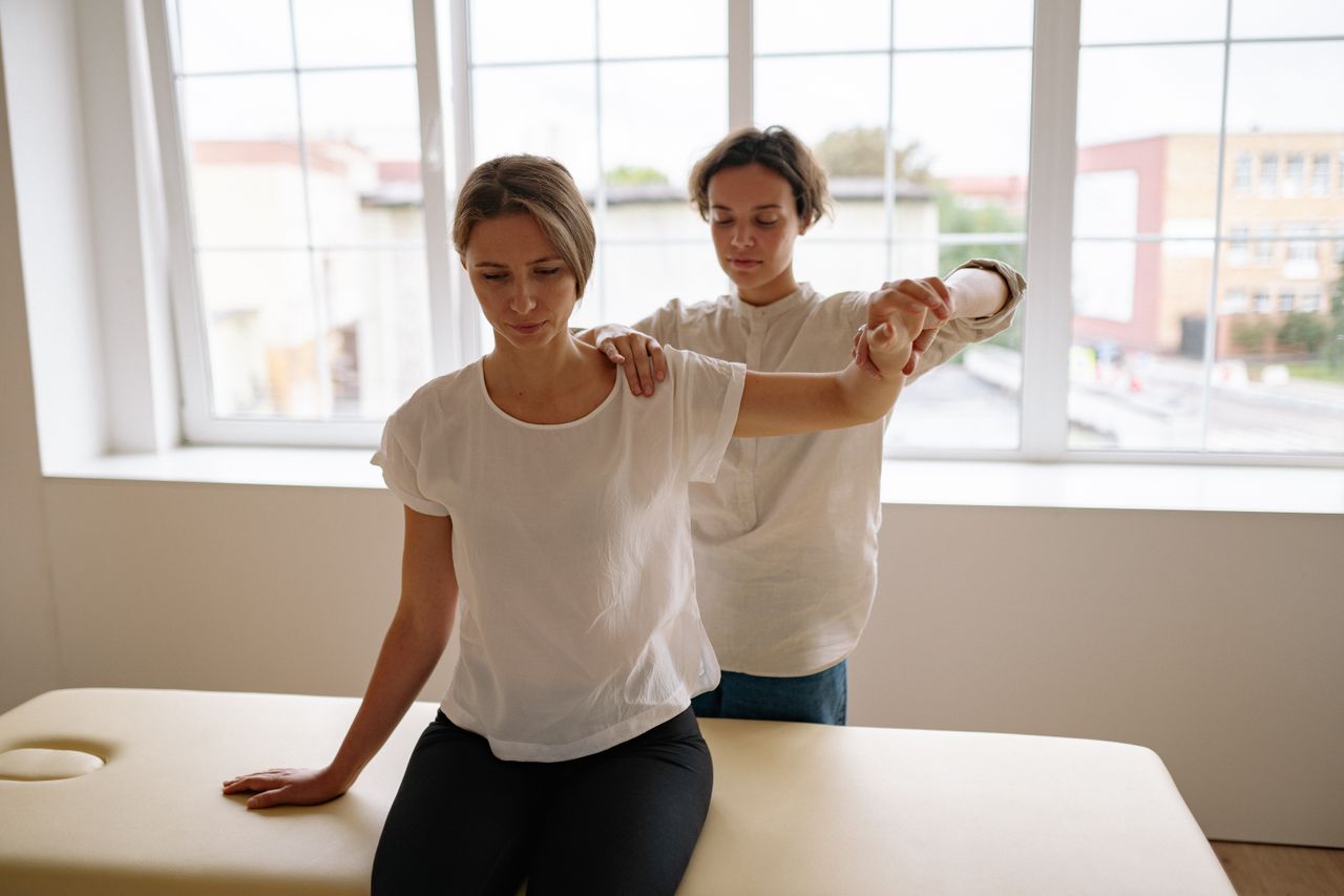 services-01 A therapist assists a woman with arm stretching during a physical therapy session in a bright, indoor setting.